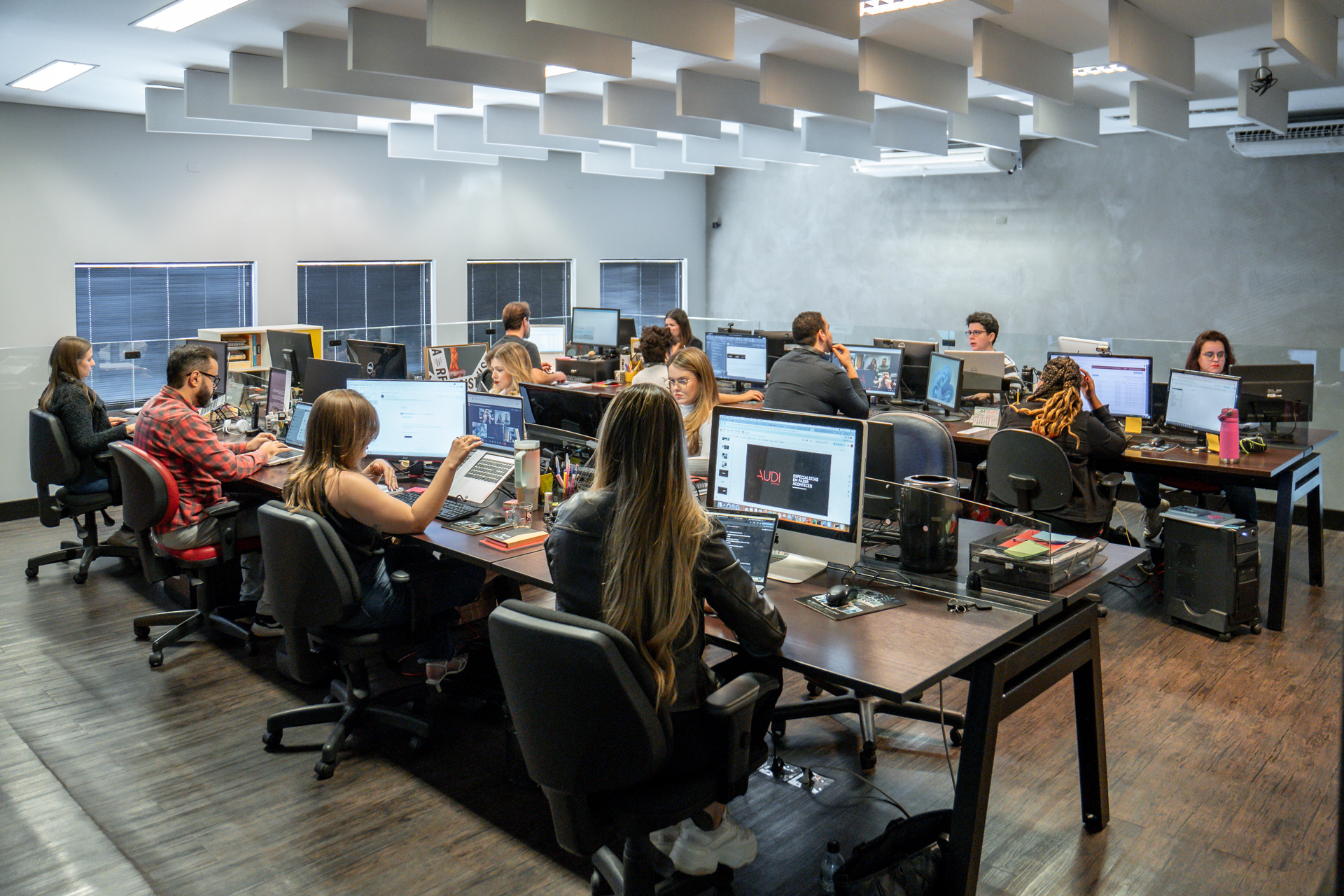 Equipe trabalhando na Audi Comunicação, com várias pessoas sentadas em mesas coletivas usando computadores.